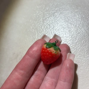 Jordgubbsberlock i plast - Supergullig berlock formad som en jordgubbe i röd och grön plast. Perfekt att hänga på ett halsband eller armband för en söt och somrig vibe. Realistisk design med små prickar och gröna blad upptill.
