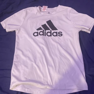 Vita Adidas t-shirts med tryck, 152 - Två vita Adidas t-shirts i klassisk modell med rund hals och kort ärm. Ena har stort svart Adidas-logga framtill, den andra har Adidas-tryck i kontur. Perfekta för sport eller chill. Materialet är mjukt och andas, troligtvis polyester.