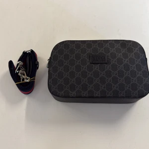 Svart axelväska från Gucci med GG-mönster - Snygg svart axelväska från Gucci med klassiskt GG-mönster över hela väskan. Väskan har en avtagbar axelrem i marinblått med röda detaljer och silverfärgade metalldetaljer. Tillverkad i slitstarkt syntetmaterial och skinn, perfekt för dig som gillar stilrena designer.