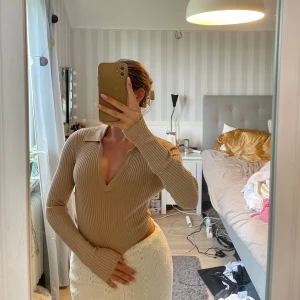 Beige ribbad stickad polotröja - Supersnygg beige stickad polotröja med djup v-ringning och krage. Tröjan är ribbad och har lång ärm, perfekt för lager-på-lager och en clean look. Sitter tajt och framhäver figuren, passar till både jeans och kjol.