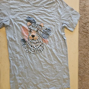Ljusblå t-shirt med leopardtryck Gill - Ljusblå t-shirt från Gill Nightwear med coolt leopardtryck framtill och tropiska blad i blått och rosa. Klassisk rund halsringning och korta ärmar. Perfekt för dig som gillar djurmönster och vill sticka ut lite extra.