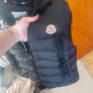 Svart Moncler dunväst med huva M - Snygg svart dunväst från Moncler med huva och tydlig logga på bröstet. Västen har två stora dragkedjefickor framtill och broderad Moncler-text på huvan. Perfekt för lager-på-lager och streetstyle. Fodrad och quiltad för extra värme.