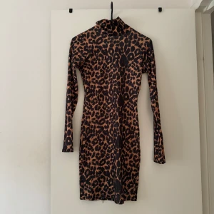 Leopardmönstrad klänning PrettyLittleThing - Snygg tight leopardmönstrad kortklänning från PrettyLittleThing med lång ärm och hög krage. Klänningen har bruna och svarta toner och är tillverkad i ett stretchigt syntetmaterial som sitter figurnära och markerar siluetten.