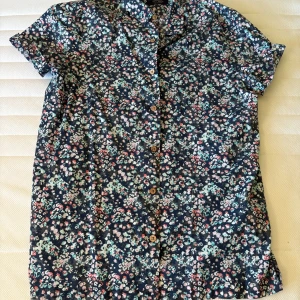 Blommig kortärmad skjorta i bomull - Snygg kortärmad skjorta med blommigt mönster i blått, grönt, rosa och vitt. Skjortan har klassisk krage och knäppning med träfärgade knappar. Perfekt för dig som gillar färg och mönster. Materialet är mjuk bomull som känns skönt mot huden.