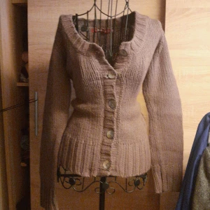 Beige stickad kofta från Indiska - Mysig beige stickad kofta från Indiska med stora knappar framtill och bred ribbad kant nedtill och vid ärmslut. Koftan har lång ärm och en djupare ringning, perfekt för lager på lager. Snygg och enkel att matcha med olika outfits.