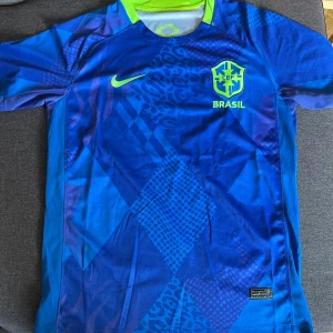 Blå Brasilien Nike fotbollströja - Snygg blå Brasilien fotbollströja från Nike med korta ärmar. Tröjan har ett unikt mönster med leoparddetaljer och geometriska former, samt neongula detaljer på logga och Nike-swoosh. Tillverkad i lätt och ventilerande polyester, perfekt för match eller träning. Det är storlek S