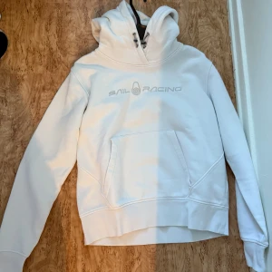 Vit hoodie från Sail Racing med huva - Äkta!!!! Vit hoodie från Sail Racing med broderad logga på bröstet. Tröjan har stor huva med snörning, magficka och ribbade muddar. Perfekt för dig som gillar en clean och sportig stil. Mjuk insida och relaxed fit. Storlek L, men tror den passar mycket bra för M samt S, äkta köpt från sail racing.