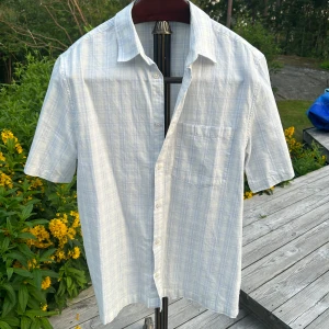 Ljusblå rutig skjorta från Weekday - Kortärmad skjorta från Weekday i ljusblått och vitt rutigt mönster. Skjortan har klassisk krage, bröstficka och knäppning framtill. Tillverkad i tunn bomull, perfekt för varma dagar. Enkel och stilren vibe. Smuts fläck på krage och höger axel men inget som inte går bort i en tvätt.