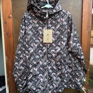 Burberry vindjacka med logomönster XL - Snygg svart vindjacka från Burberry i storlek XL med all-over logomönster i vitt, grått och röda detaljer. Jackan har huva, hel dragkedja och är tillverkad i lätt polyester. Perfekt för dig som vill sticka ut med en exklusiv streetstyle-look.