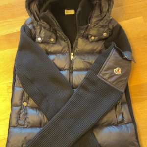 Svart Moncler cardigan med huva - Säljer en blå cardigan S/M från Moncler med avtagbar huva och ribbstickade ärmar. Jackan har dragkedja framtill, två fickor med tryckknappar och Moncler-logga på ärmen. Kombinerar dunfylld kropp med stickade detaljer för en riktigt clean look.