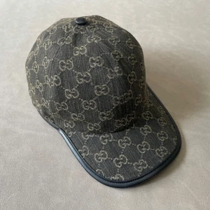 Svart keps från Gucci  - Snygg svart keps från Gucci i jeansmaterial med klassiskt GG-monogram över hela kepsen. Böjd skärm med svart kant i skinn och justerbar rem bak. Perfekt för dig som vill ha en stilren och lyxig accessoar med streetkänsla. Helt ny Aldrig använd. Pris kan diskuteras.