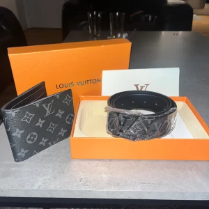 Louis Vuitton svart monogram bälte - Snyggt svart bälte från Louis Vuitton med klassiskt monogrammönster i grått och silver. Bältet är gjort i exklusivt skinn och har en stor LV-logga som spänne. Perfekt accessoar för att lyfta din outfit med lyxig känsla.
