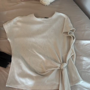 Beige topp med knyt från Zara - Snygg beige topp från Zara med korta ärmar och knyt i sidan. Toppen har en enkel, stilren design och är tillverkad i ett mjukt, stickat material som känns skönt mot huden. Perfekt för dig som gillar minimalistisk stil med en twist.