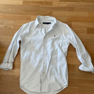 Vit skjorta från Ralph Lauren - Klassisk vit skjorta från Ralph Lauren med broderad logga på bröstet. Skjortan har lång ärm, knappar framtill och en snygg krage. Tillverkad i bomull och passar perfekt till en clean och stilren look.