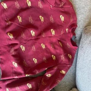 Vinröd Nike sweatshirt med logga - Säljer en vinröd sweatshirt från Nike i storlek S. Tröjan har all-over print med Nike-loggor i gult och klassisk rund hals. Mjuk och skön bomullsmix, perfekt för chill dagar eller streetstyle. Långärmad och normal passform.