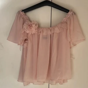 Ljusrosa blus med volang och blomdetalj  - Supersöt ljusrosa blus med volang, jättegullig rosa blomma som detalj🩷 testad 1 gång och säljer den pga att den inte passar mej i storlek, köpt i storleken XS men skulle säga att den passar S mer. Kostar 549 egentligen 💕