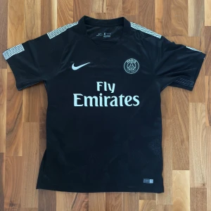 Svart PSG fotbollströja med mönster - Tja, säljer min svarta Paris Saint-Germain fotbollströja från Nike. Tröjan har korta ärmar, diskret mönster i tyget (se på bild) och vita detaljer på axlarna. Tröjan är i storlek S men kan även passa M. Den är i nytt skick och vid funderingar är det bara att höra av sig!