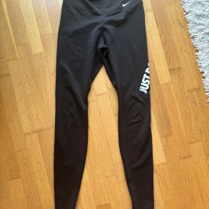 Nike Dri-Fit svarta leggings - Svarta Nike Dri-Fit leggings med trycket 'JUST DO IT' längs ena benet. Tight passform och tillverkade i ett stretchigt syntetmaterial som andas, perfekt för träning. Klassisk Nike-logga vid midjan och hög komfort.