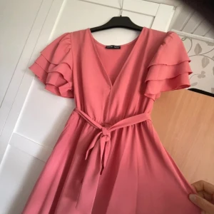 Rosa omlottklänning från SHEIN - Supersöt rosa klänning från SHEIN med volangärmar, v-ringning och knytband i midjan. Klänningen har en omlott-look och är i en lätt, flowy modell som ger en romantisk vibe. Perfekt för dig som gillar feminina detaljer och vill sticka ut lite extra.