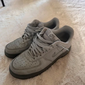 Grå Nike Air Force 1 sneakers - Snygga grå Nike Air Force 1 sneakers med klassisk siluett och lågt skaft. Skorna har ovandel i mocka, perforerad tå och svart sula. Ikonisk Swoosh-logga på sidan och AIR-detalj på sulan. Perfekta för dig som gillar streetstyle och vill ha en clean look.