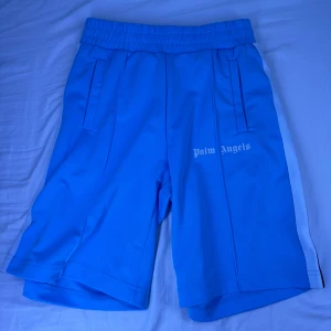 Blå Palm Angels shorts (oanvända) - (Pris kan diskuteras) Snygga blå shorts från Palm Angels med vit sidostripes och elastisk midja. Klassisk sportig look med två fickor fram och logga tryckt på ena benet. Perfekta för sommaren och chill dagar.