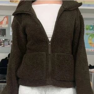 Mörkgrön teddyjacka med dragkedja - Mysig mörkgrön teddyjacka med stora fickor framtill och dragkedja hela vägen upp. Jackan har en bred krage och är långärmad, perfekt för kyliga dagar. Den har en loose passform och är tillverkad i fluffigt teddy-material som ger en chill vibe.