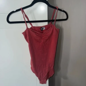 VinRöd body från H&M - Säljer en röd bodysuit från H&M med smala axelband och rynkdetalj framtill. Perfekt att styla med jeans eller kjol för en snygg look. Mjuk och stretchig bomullskänsla som sitter tight och bekvämt.