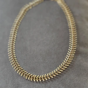 Guldigt kedjehalsband med fiskbensmönster - Snyggt halsband i guldton med fiskbensinspirerat mönster. Kedjan har en justerbar längd och stängs med en karbinhake. Perfekt för att ge din outfit en cool och trendig touch.