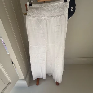 Vit långkjol från Gina Tricot - Säljer en vit långkjol från Gina Tricot med smock i midjan. Kjolen är luftig och har en lätt, flowy känsla som passar perfekt till varma dagar. Tillverkad i mjukt bomullstyg och har en enkel, stilren look.