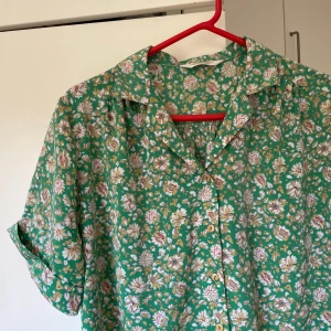 Grön blommig skjorta med kort ärm - Snygg grön skjorta med korta ärmar och blommigt mönster i vitt, gult och rosa. Skjortan har klassisk krage och knäppning framtill med ljusa knappar. Oversized 
