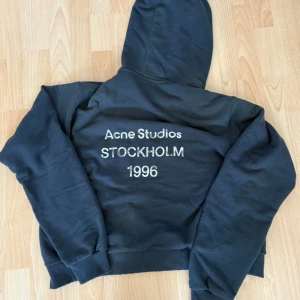 Svart hoodie Acne Studios S - Svart hoodie från Acne Studios med tryck på ryggen: 'Acne Studios STOCKHOLM 1996'. Klassisk passform med huva och muddar vid ärmslut och midja. Tillverkad i mjuk bomullsblandning med 11% polyester. Perfekt för en chill och stilren look.