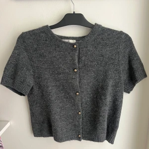 Grå stickad kofta från H&M - Mysig grå stickad kofta från H&M med korta ärmar och snygga guldfärgade knappar framtill. Koftan har rund halsringning och ribbade kanter för en clean look. Perfekt att slänga över en topp för en chill vibe.