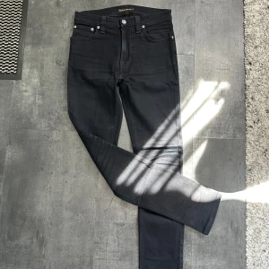 Svarta  jeans från Nudie Jeans - Säljer ett par svarta jeans från Nudie Jeans i modellen Tight Long John. Jeansen har klassisk femficksdesign, svarta sömmar och Nudie-logga på bakfickan. Tillverkade i stretchig bomullsdenim för en tight passform.