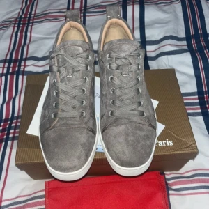  Christian Louboutins - Snygga grå sneakers i mocka från Christian Louboutin med klassisk röd sula och vita kanter. Skorna har rund tå, snörning och diskreta detaljer i beige läder på insidan. Perfekta för dig som vill sticka ut med exklusiv streetstyle.