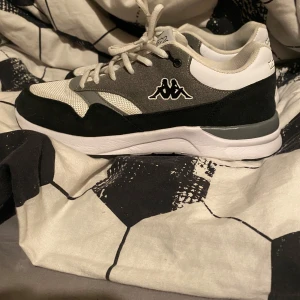Svarta och vita sneakers från Kappa - Snygga sneakers från Kappa i svart, vitt och grått med klassisk logga på sidan. Ovandelen är i mesh och mocka med vita snören och en bekväm, tjock vit sula. Perfekta för dig som gillar sportig och stilren look. Helt ny, inte använd!!