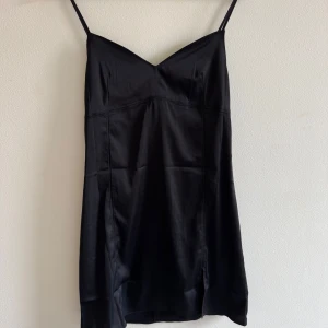 Svart satin slipklänning med slits - Snygg svart slipklänning i satin med smala axelband och v-ringad hals. Klänningen har en liten slits framtill och sömmar som ger en cool, figurnära siluett. Perfekt för dig som gillar minimalistisk stil och vill ha något enkelt men ändå elegant.
