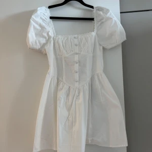 Vit kort klänning från Boohoo - Supersöt vit klänning från Boohoo med puffärmar och knappar framtill. Klänningen har en fyrkantig ringning med spetskant och är tillverkad i bomull. Passformen är normal och kjolen är lätt utställd. Prislappen är kvar💓