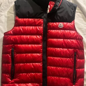 Röd dunväst från Moncler - Snygg röd dunväst från Moncler med svart axelparti och Moncler-logga på bröstet. Västen har quiltad design, två dragkedjeförsedda fickor och en svart dragkedja med Moncler-tryck. Perfekt för att lyfta din outfit under kyliga dagar.