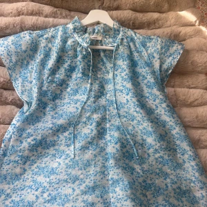 Blommig blå kortklänning från Samsøe - Supersöt blus från Samsøe Samsøe med ljusblått blommigt mönster på vit botten. Blusen har korta ärmar, knytband vid halsen och en loose passform. Materialet har en lätt glansig yta som ger en fräsch vibe. Perfekt för varma dagar!💙🤍