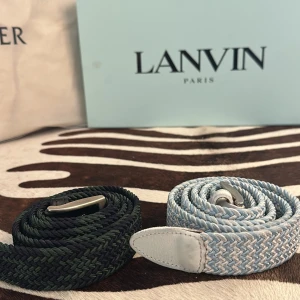 Två flätade bälten  - Säljer två snygga flätade bälten från Lanvin. Ett är ljusblått och vitt med vita skinn-detaljer, det andra är mörkgrönt och svart. Båda har silverfärgade spännen och är perfekta för att lyfta din outfit med en stilren touch.