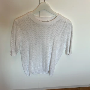 Vit stickad topp - Säljer en vit stickad topp med korta ärmar och snyggt hålmönster . Rund halsringning med bred ribbad kant och mjukt material som känns skönt mot huden.