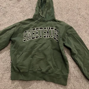Grön SWEETSKTBS hoodie med huva - Säljer en grön hoodie från SWEETSKTBS med stor vit och svart logga på bröstet. Tröjan har huva med snörning, magficka och ribbade muddar. Perfekt för chill dagar och streetwear-stil. Materialet är mjuk bomull och passformen är normal. Den har två små hål i bak men inget man tänker på. De syns på sista bilden