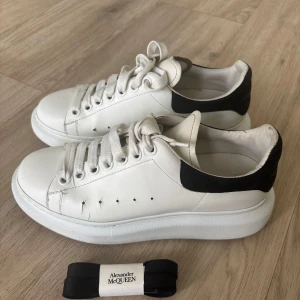 INTRESSEKOLL Alexander McQueen vita sneakers - INTRESSEKOLL på dessa stilrena vita sneakers från Alexander McQueen med svart hälparti i mocka och tjock platt sula. Skorna har snörning och rund tå, samt diskret logga baktill. Medföljer extra svarta skosnören och dustbag.