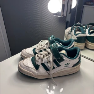 Adidas Forum Low vita och gröna sneakers - Snygga Adidas Forum Low sneakers i vitt och mörkgrönt skinn med klassiska tre ränder på sidan. Skorna har snörning och kardborreband över vristen. Nypris 1145kr💓