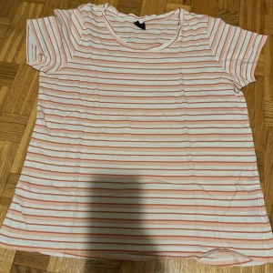 Randig vit och rosa t-shirt i bomull - Fräsch t-shirt med vita och ljusrosa horisontella ränder. Modellen har rund hals och korta ärmar, perfekt för chill dagar. Materialet är mjuk bomull som känns skönt mot huden. Passformen är normal och t-shirten har en enkel, clean vibe.