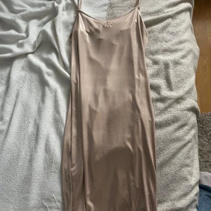 Champagnefärgad slipklänning Calvin Klein - Säljer en champagnefärgad slipklänning från Calvin Klein Jeans med tunna axelband och broderad CK-logga framtill. Klänningen har en glansig finish och är gjord i ett mjukt, slätt material som ger en lyxig känsla. Perfekt för dig som gillar minimalistisk och stilren design.