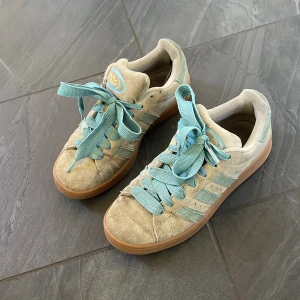 Adidas Campus sneakers i beige och blå - Adidas Campus sneakers med beige mocka och ljusblå detaljer, inklusive de klassiska tre ränderna och snörning. Skorna har en brun gummisula och guldfärgat Adidas-logo på hälen. Perfekt för dig som gillar retrovibe och snygga färgkombinationer.