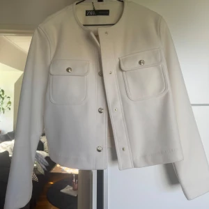 Vit kavajjacka från Zara, M - Snygg vit kavajjacka från Zara med rund halsringning och guldiga knappar. Jackan har två stora bröstfickor med lock och är i en rak, lite boxig modell. Perfekt att slänga över en topp för en clean look.