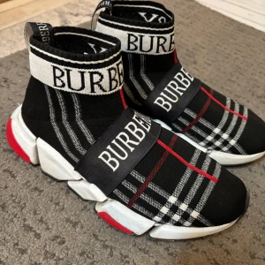 Burberry skor - Burberry sock sneakers i svart med vit och röd rutig design, tydlig logga på resåren och ovandelen. Skorna har chunky vit och svart sula med röda detaljer. Stickad ovandel i syntetmaterial ger en tight och sportig känsla.