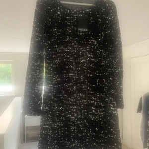 Mörkgrå glittrig paljettklänning Pieces - Kort klänning från Pieces, täckt med glittrande paljetter som ger en riktigt festlig vibe. Klänningen har lång ärm och rund halsringning, perfekt för att sticka ut på nästa event. Materialet är syntet och passformen är normal.